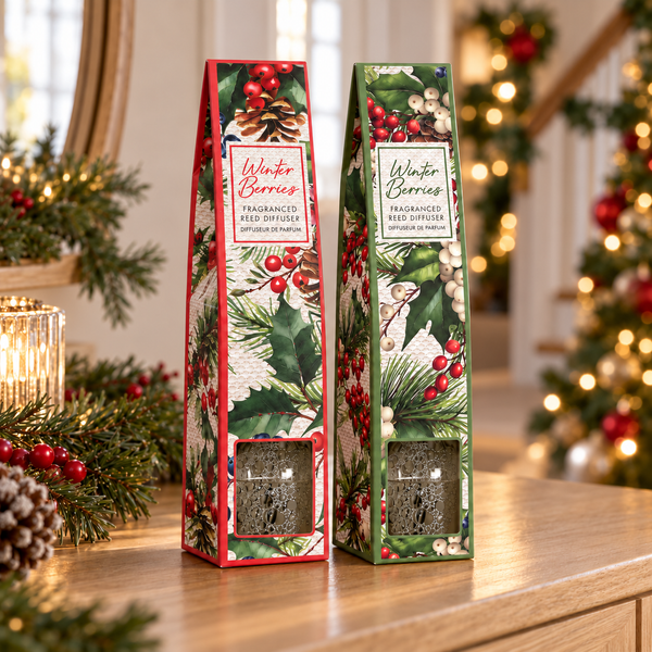 Two tall, narrow Winter Berries fragranced seed diffuser boxes standing on a wooden table in a festive holiday setting. The packaging features a red border on the left box and a green border on the right box, both decorated with illustrations of winter berries, pine needles, and snowy white elements.