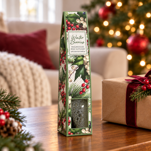 A packaged Winter Berries fragranced Reed Diffuser sits on a wooden table in a cozy holiday setting, with a blurred Christmas tree and warm lights in the background and a wrapped gift box beside it.