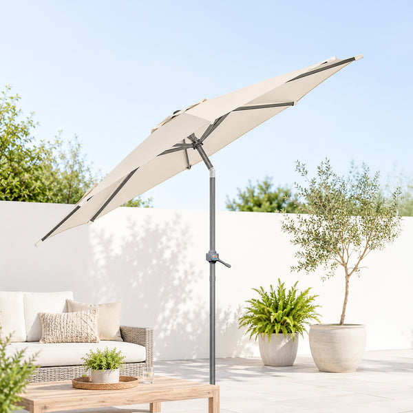 A large beige patio umbrella with a gray pole and crank handle, shown in a sunlit outdoor patio setting next to a white wicker sofa and potted plants.