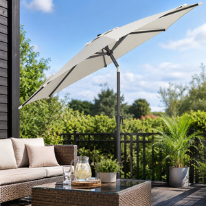 A large patio umbrella in a light beige/cream shade with a dark gray pole, shown on a wooden outdoor deck next to a wicker sofa and coffee table, with green garden foliage and a blue sky in the background.