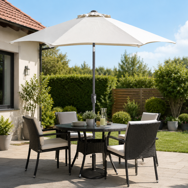 Outdoor patio dining set with a round dark gray wicker table, four matching armchairs with light beige cushions, and a large cream patio umbrella on a paved patio in front of a house.