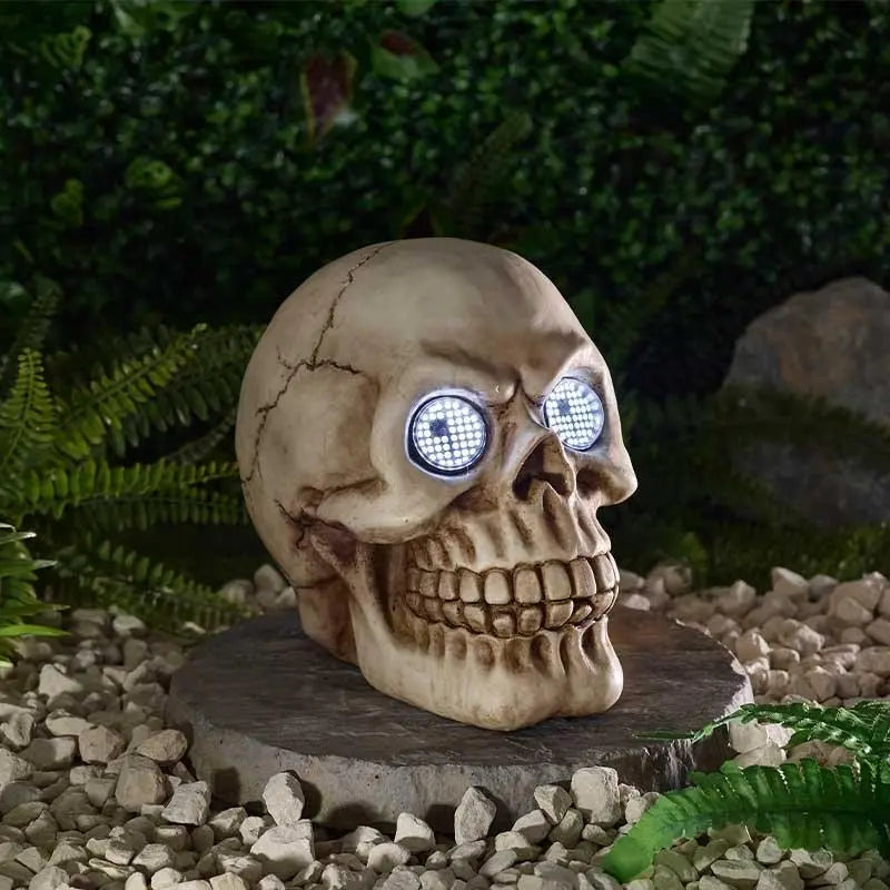 A decorative skull garden statue with glowing blue LED eyes, displayed on a stone base outdoors among green foliage and white gravel.