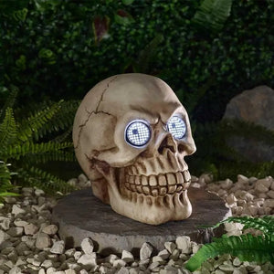 A decorative skull garden statue with glowing blue LED eyes, displayed on a stone base outdoors among green foliage and white gravel.