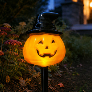 A Halloween yard light shaped like a glowing jack-o'-lantern pumpkin wearing a black witch hat on a black stake, photographed outdoors in a garden at night.