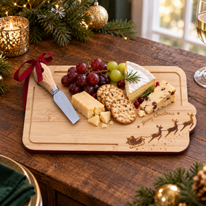A rectangular wooden charcuterie board with a holiday engraving of Santa in a sleigh and the words 'Merry Christmas,' styled on a dark wooden table with cheese, grapes, crackers, and a small knife. Christmas greenery, lights, ornaments, and a candle are visible in the background.