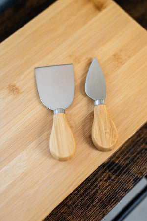 Two cheese knives with wooden handles on a bamboo cutting board.