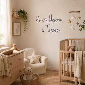 Black metal script wall art reading 'Once Upon a Time' mounted on a beige wall in a nursery between a wooden cot, a shelf with a plant and bunny on it, soft natural colours