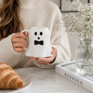 A person in a cream knit sweater holds a white ghost-shaped mug with a black face and bow tie. The mug is shown on a marble table beside a croissant, a glass vase with baby's breath, and a book titled KINFOLK.