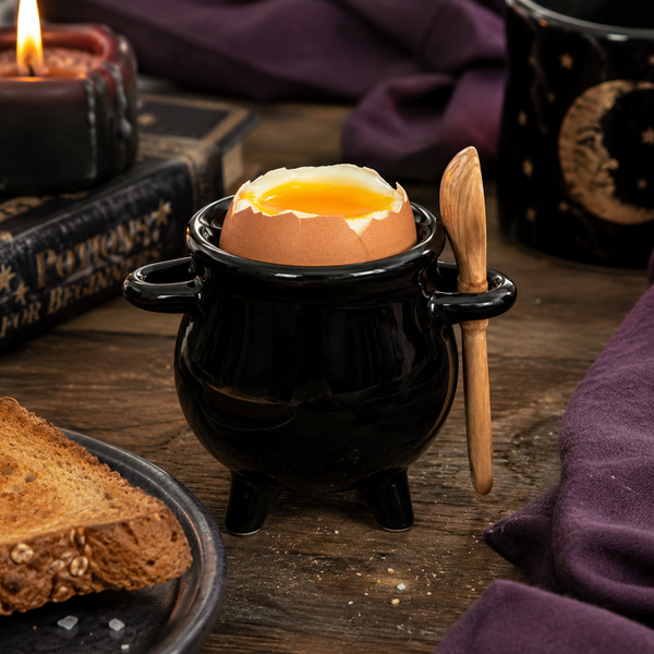 A black cauldron-shaped egg holder with small handles and legs, holding a cracked egg with a soft yolk, alongside a wooden spoon. The scene is styled on a rustic wooden table with a slice of toast, a lit candle, old books, and purple fabric in the background.