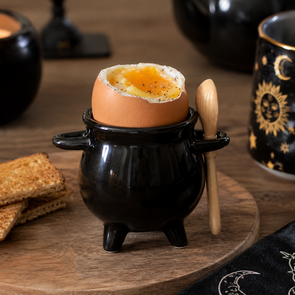 A black ceramic cauldron-shaped egg holder with a halved hard-boiled egg inside, a small wooden spoon resting on the side, and toasted bread on a wooden board in a cozy tabletop setting with a teapot and candle in the background.