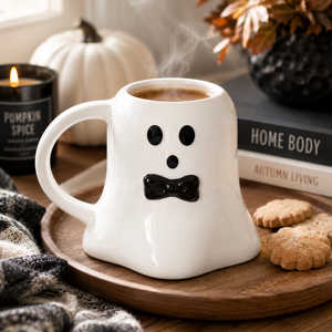 A white ceramic mug shaped like a ghost with a black face and bow tie, filled with a hot beverage and shown on a wooden tray in a styled autumn setting with cookies, a candle, and books.