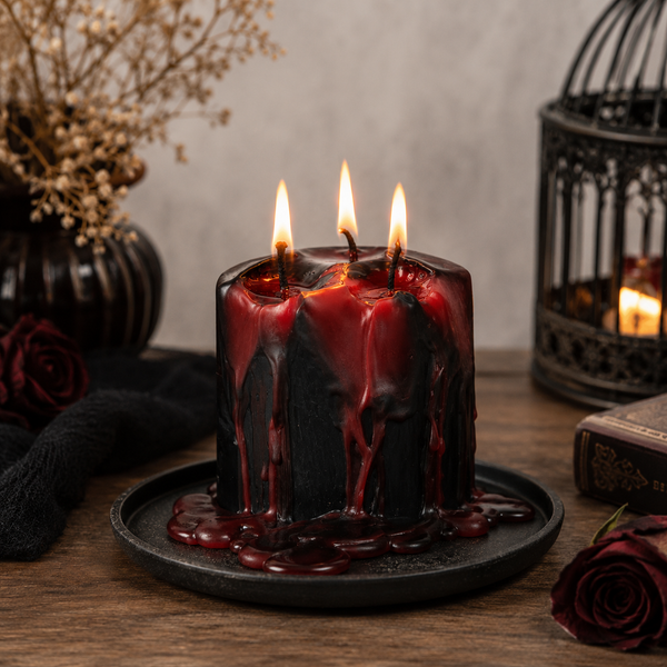 A black pillar candle with dramatic red wax drips and three lit wicks, displayed on a dark plate on a wooden table with gothic-themed props including dried flowers, a black birdcage, and dark roses.