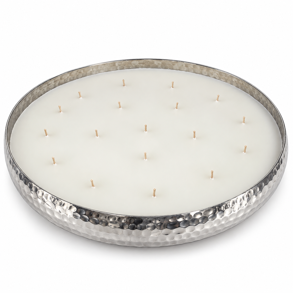 A large round wax candle in a hammered metal bowl, lit with multiple small flames, set on stone piece, in a bright and white hallway.
