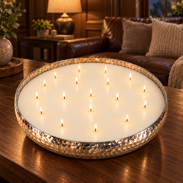 A large round wax candle in a hammered metal bowl, lit with multiple small flames, set on a wooden table in a cozy living room with a leather sofa in the background.