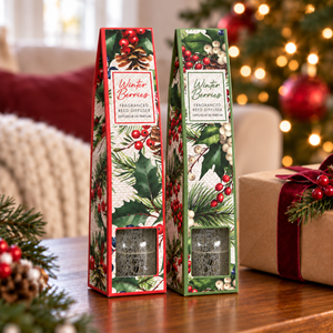 A packaged Winter Berries fragranced Reed Diffuser sits on a wooden table in a cozy holiday setting, with a blurred Christmas tree and warm lights in the background and a wrapped gift box beside it.