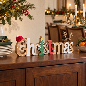 Wooden decorative letters spelling Christmas arranged on a dark wood cabinet in a warmly lit holiday dining room. The letters include a tree, star, reindeer antlers, and a small red hat, with a blurred background of candles and tables.