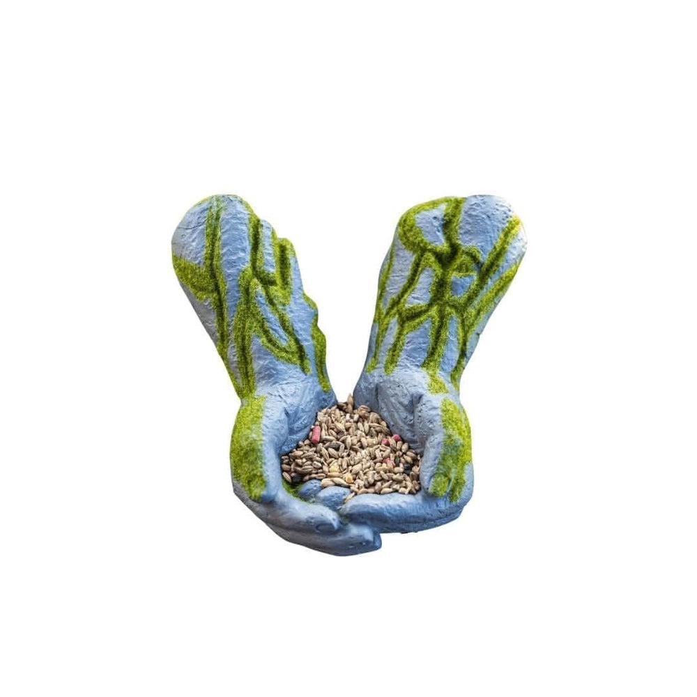 A garden-style hand-shaped planter or seed holder made of textured gray material with green mossy detailing, holding a pile of birdseed on a bright green grass lawn.