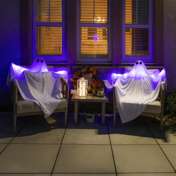 Two ghost-shaped outdoor decorations on chairs with a small table between them, illuminated by purple and white lights.