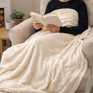 A person sitting in a beige armchair, wrapped in a thick cream-colored plush blanket and reading an open book. The scene is set in a cozy living room with a small wooden side table, a white mug, and a potted plant in the background.