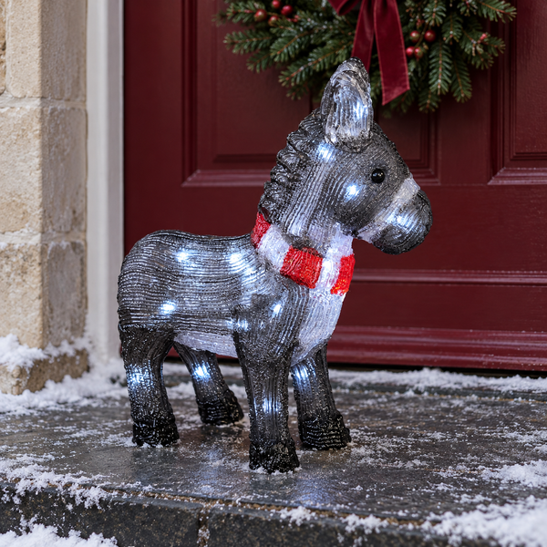 A lit decorative donkey figure with a red and white striped scarf stands on a snowy doorstep in front of a red door and holiday wreath.