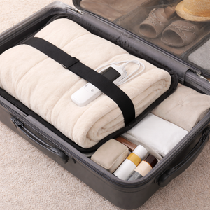 A folded cream-colored blanket or quilt packed inside an open black suitcase, secured with a black strap and a white electric heating pad. Other packed items like shoes, a hat, and toiletries are visible in the suitcase compartments.