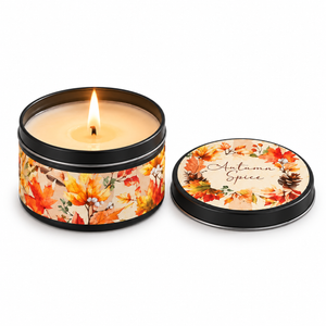 A lit candle in a round black metal tin with a warm autumn leaf print, shown next to its matching lid on a white background. The lid features the scent name 'Autumn Spice' in script.