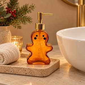 A gingerbread-shaped amber glass soap dispenser with a gold pump and handle, styled on a stone tray next to a white sink bowl, a rolled towel, and holiday greenery in a warm bathroom setting.