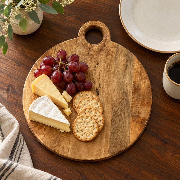 Round wooden serving board with a handle, styled with cheese wedges, red grapes, and crackers on a dark wood table, with a plate and cup nearby.