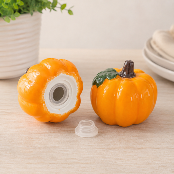 An orange pumpkin-shaped ceramic container shown open on a light wood surface, with its white interior visible and a small detached lid in front. A second closed pumpkin container stands to the right with a green leaf detail and dark stem.