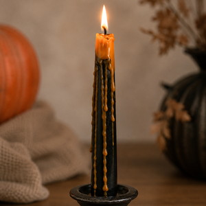A black taper candle with orange wax dripping down the sides, lit and standing in a dark metal holder on a wooden surface, with a pumpkin and dried leaves in the softly blurred background.