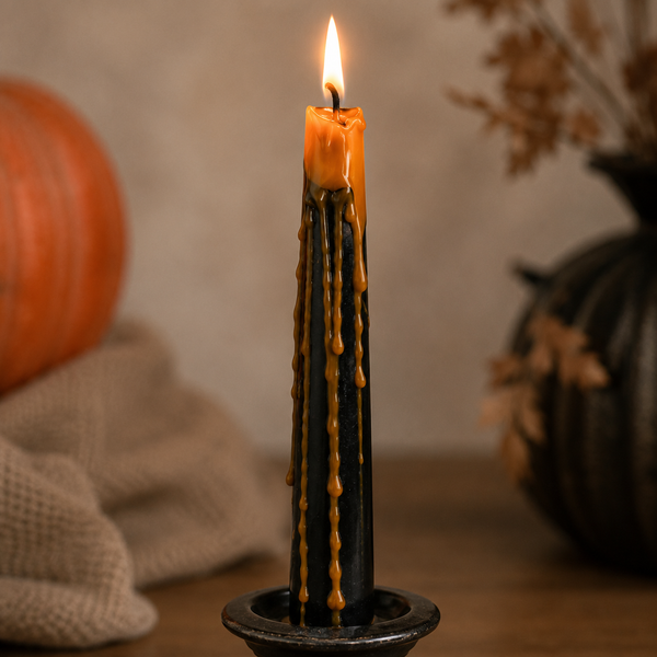 A black taper candle with orange wax dripping down the sides, lit and standing in a dark metal holder on a wooden surface, with a pumpkin and dried leaves in the softly blurred background.