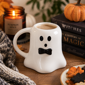 A white ceramic mug shaped like a ghost with a black face and bow tie, filled with coffee, styled on a wooden table with a candle, books, a pumpkin, and a knit blanket in a cozy autumn setting.