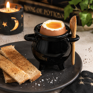 A black ceramic cauldron-shaped mug holding a soft-boiled egg with a wooden spoon resting inside, set on a dark plate with toasted bread, a lit candle, and a book in the background.