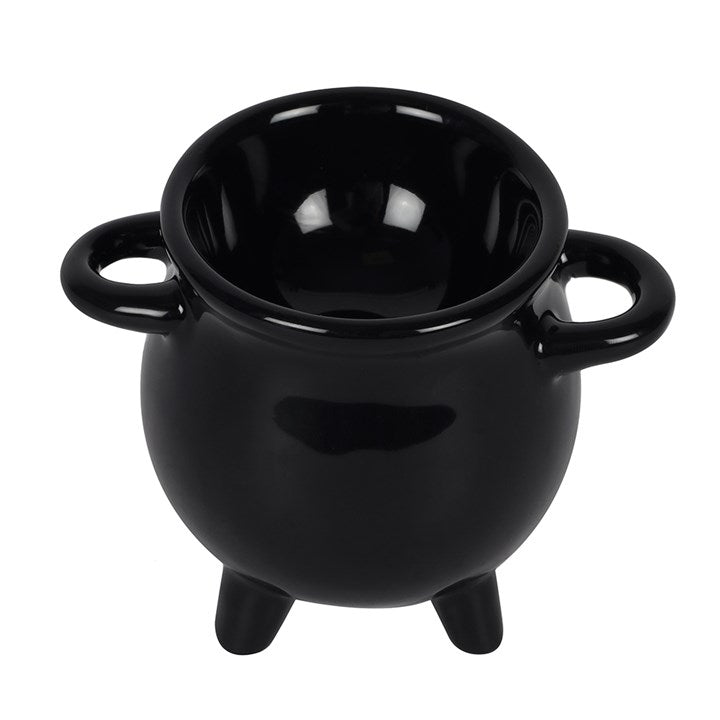 A black ceramic cauldron-shaped egg holder with a halved hard-boiled egg inside, a small wooden spoon resting on the side, and toasted bread on a wooden board in a cozy tabletop setting with a teapot and candle in the background.