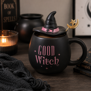 A black ceramic mug with a lid shaped like a witch hat, decorated with pink 'GOOD WITCH' text and a gold crescent moon and star spoon, styled on a wooden table with candles and books.