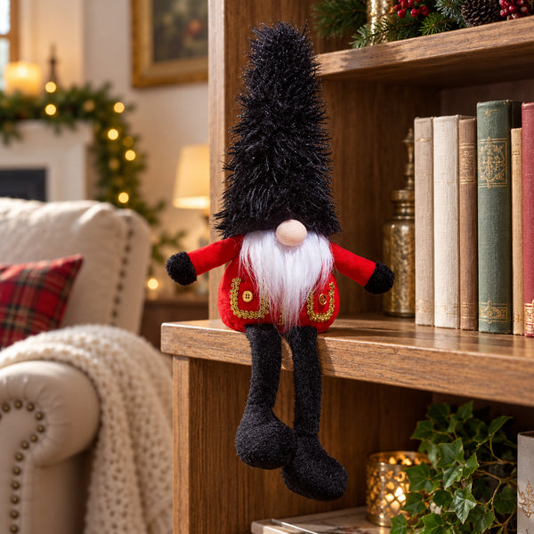 A Christmas gnome plush decoration with a tall black shaggy hat, white beard, red coat with gold trim, and black legs, posed on a wooden bookshelf in a cozy living room setting.