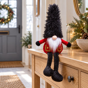 Christmas guard gonk with a tall black hat, red coat and dangling legs, sitting on an oak hallway table beside festive greenery.