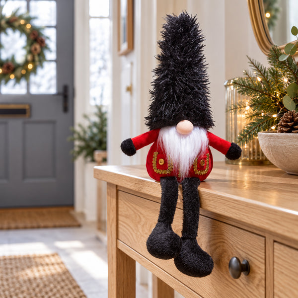 Christmas guard gonk with a tall black hat, red coat and dangling legs, sitting on an oak hallway table beside festive greenery.