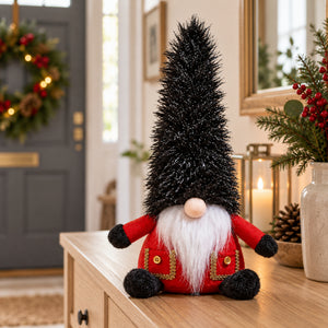 A Christmas gnome decoration with a tall black glittery hat, red coat with gold trim, white beard, and black pom-pom hands, displayed on a wooden table in a decorated home interior.