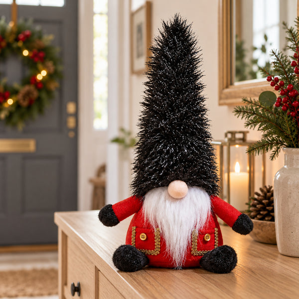 A Christmas gnome decoration with a tall black glittery hat, red coat with gold trim, white beard, and black pom-pom hands, displayed on a wooden table in a decorated home interior.