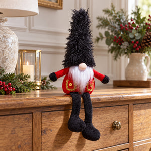 Christmas guard gonk with a tall black hat, red coat and dangling legs, sitting on a wooden sideboard beside festive foliage.