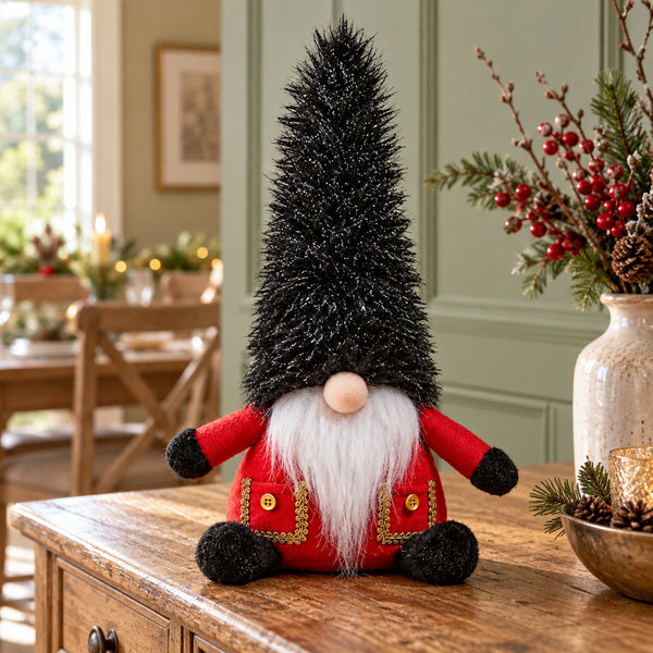 A festive Christmas gnome decoration sitting on a wooden table in a decorated dining room. The gnome features a tall black glittery cone hat, a white beard, a red body, and red pockets with gold trim.