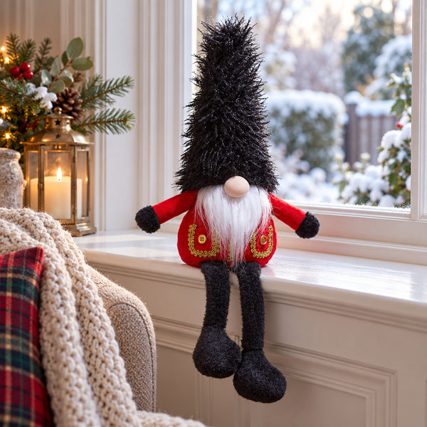 A festive Christmas gnome doll with a tall black spiky hat, white beard, red coat with gold trim, and black legs, sitting on a white windowsill in a decorated holiday room.