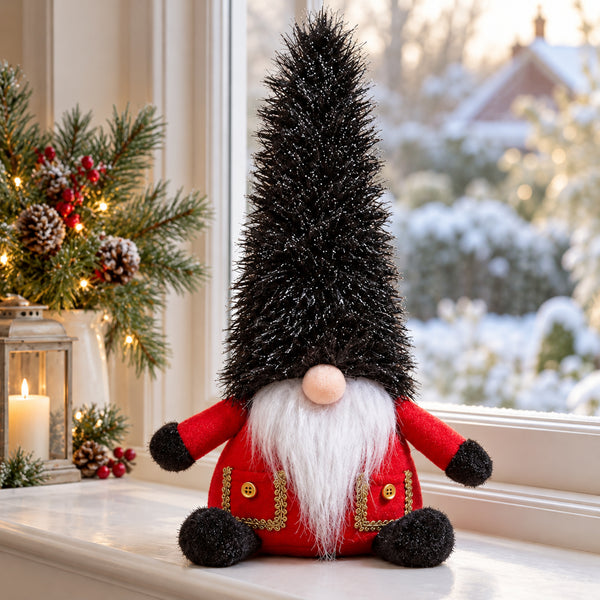A Christmas gnome decoration sitting on a windowsill, with a tall black shaggy hat, white beard, red body, and gold trim, set against a softly blurred winter window view and holiday greenery.
