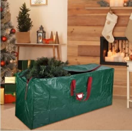 A green Christmas tree storage bag with red handles and a zipper, placed on the floor beside a decorated Christmas tree and a fireplace.