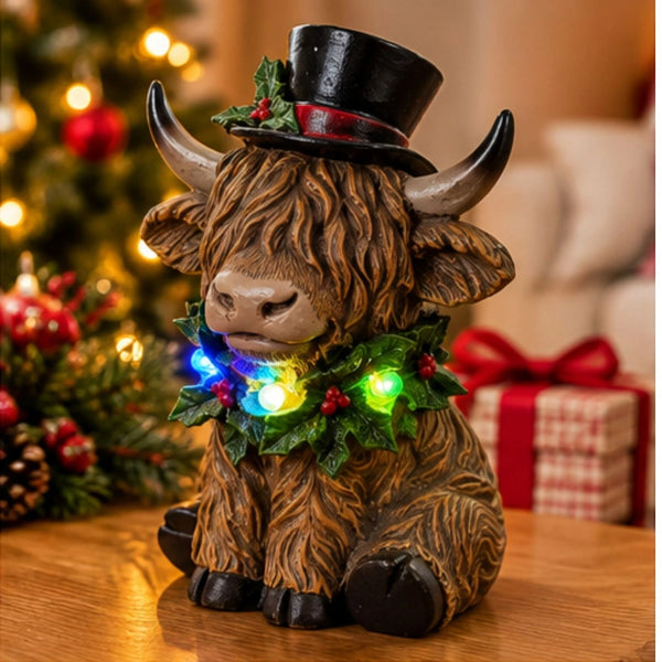 A decorative Highland cow figurine with a black top hat and a green holly garland around its neck, lit with colorful LED lights, set on a wooden table with a blurred Christmas tree and gift box in the background.