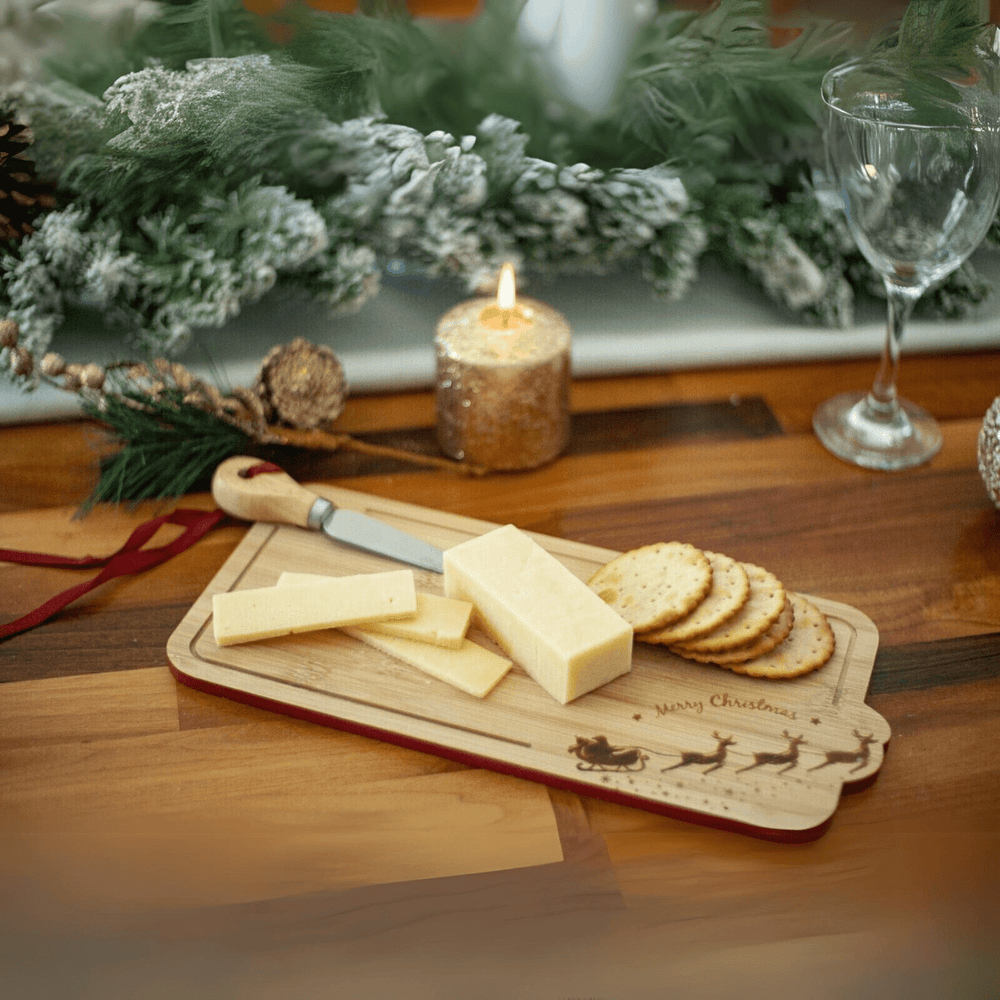 A Christmas-themed wooden cheese board with a Santa sleigh design, displayed on a table with cheese and crackers, a knife, and holiday decorations in the background.