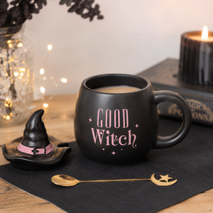 A matte black ceramic mug with 'GOOD Witch' printed in pink lettering, styled on a black cloth with a small witch hat saucer, a gold spoon, and a candle in the background.