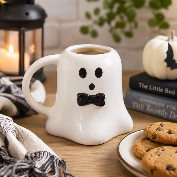 A white ceramic coffee mug shaped like a friendly ghost with a black bow-tie mouth and black eyes, filled with coffee on a wooden table surrounded by seasonal props including cookies, a lantern, and a small pumpkin.