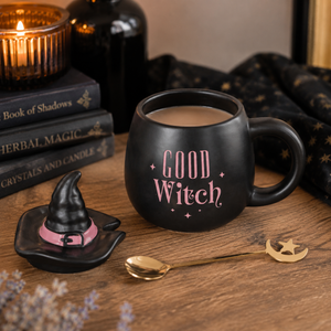 A black ceramic mug with pink 'GOOD WITCH' lettering sits on a wooden table, alongside a small witch hat saucer, a gold spoon, and a black starry cloth. Lit candles and books are blurred in the background.